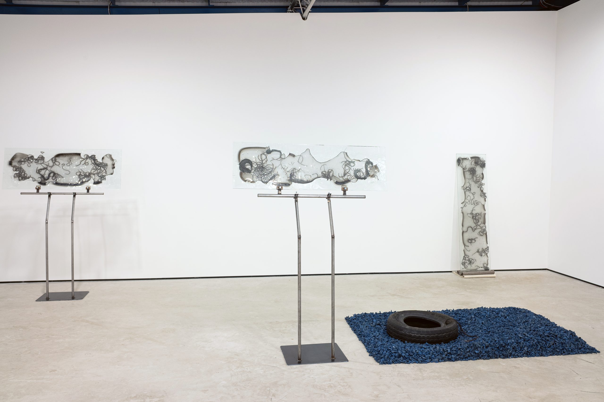 Three glass sculptures in a row, on the left two small panels propped up on metal stands and on the right a longer pane leaning against the wall. In the foreground a rectangle of blue on the floor with an old car tyre laid atop it.