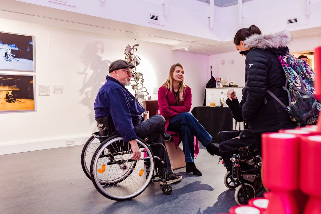 Heaton sits in his wheelchair at the centre of the room talking to a group of people.