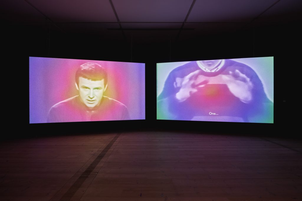 Two screens side by side. On the left screen a male figure, from the chest up. He has short hair cut in a 1960'd style. On the right a chest of a figure wearing a jumper, their hands moving towards the camera. Both images seem to be black and white but have a kind of wash or filter that is coloured, orange to purple.
