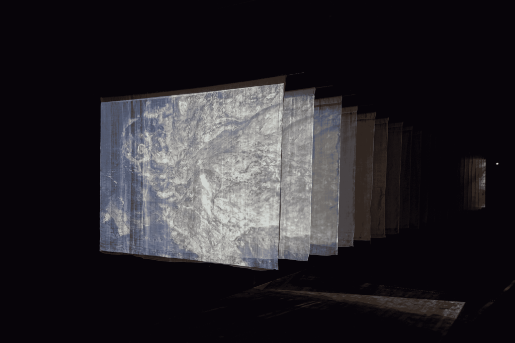 Several sheets lined up, like pages. hanging from the ceiling. The room is dark except for bright whitish imagery projected on the sheet-screens. The projection fades as it passes through each sheet.