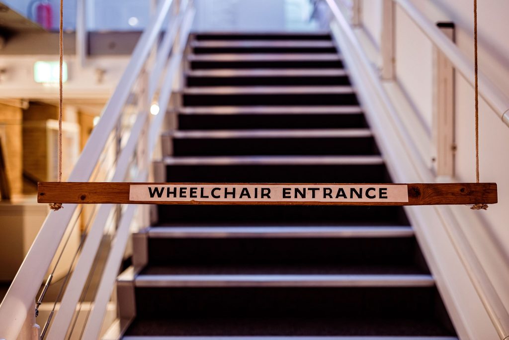 A piece of wood hangs horizontally across a staircase with the text WHEELCHAIR ENTRANCE printed in block capitals on it