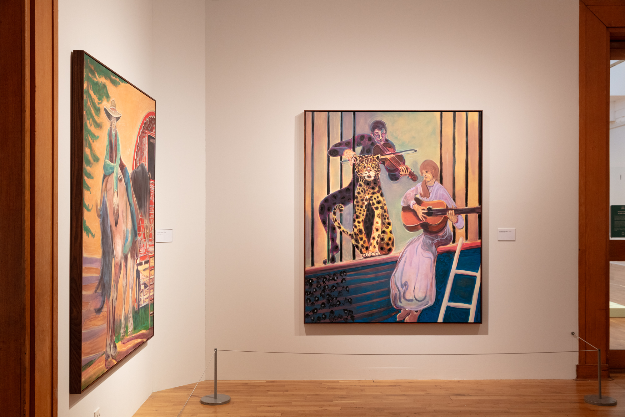 Two paintings on a gallery wall - one visible obliquely on the lefthand wall shows a person on horseback; the more central, front-on painting shows a pair of musicians and a leopard at a balcony.