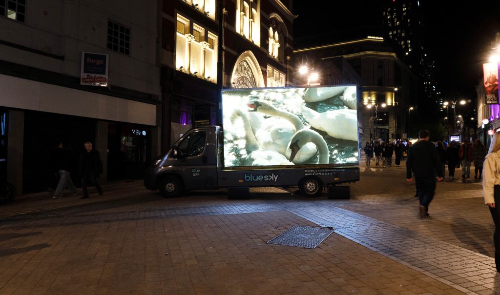 A lorry with a large screen on the back shows a film work containing swans on a busy shopping street at night