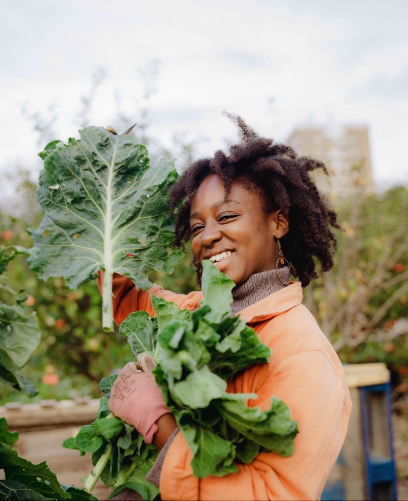 A person in a garden smiles at the camera, holding up large leaves.
