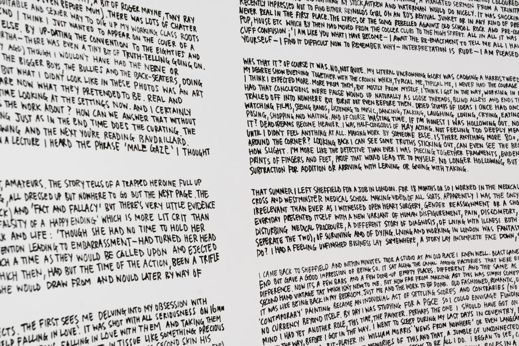 Paragraphs of black text handwritten in large block capitals written out on a white gallery wall.
