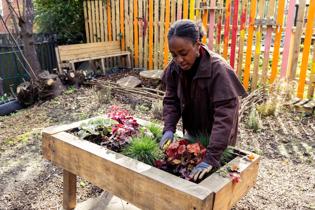 A person in long brown coat and gloves plants out a raised planter in front of a brightly coloured fence
