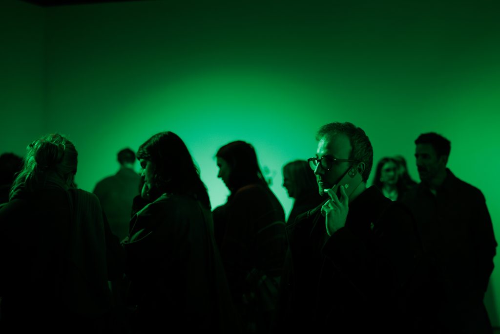 A mad holds an iPhone to his ear in a crowded room - he is listening to an audio work by the artist, surrounded in a pool of fluorescent green light