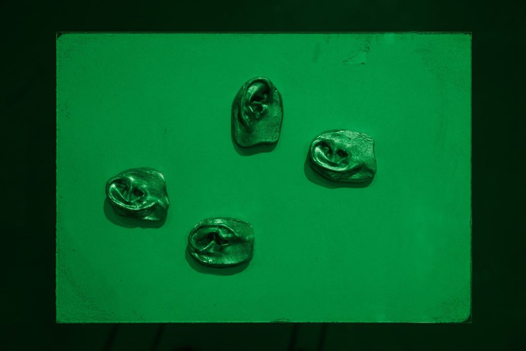 4 metallic ear-shaped sculptures lay flat on a plinth in a quadrant formation, dimly lit in green light
