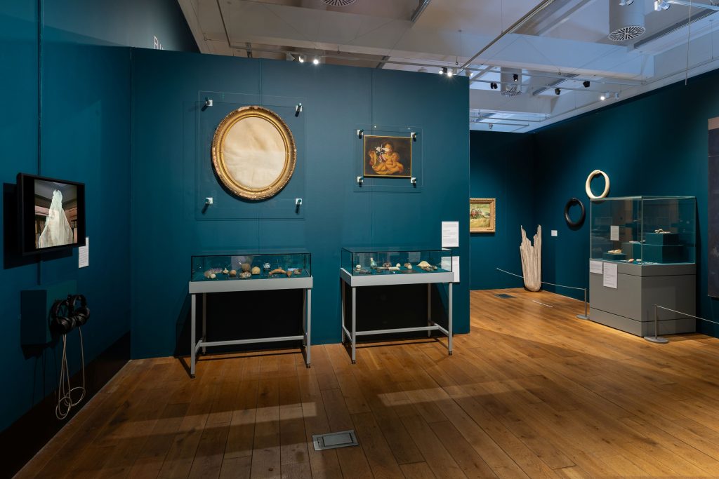 A blue-walled museum gallery. A jutting wall on the left has two vitrines pushed up against it with two painting hung above, one faded badly. In a corner beyond the jutting wall, a number of sculptural objects, including a black and a white ouroboros.