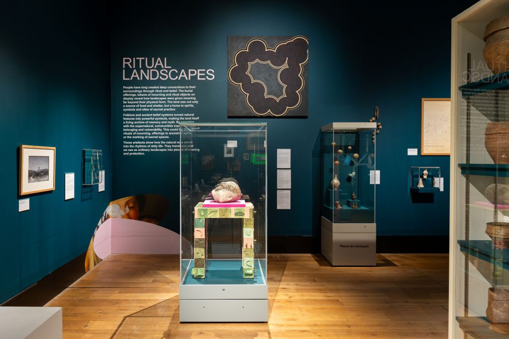 A blue-walled museum gallery. On the back wall, a vitrine containing a variety of objects and a vinyl wall text titled 'RITUAL LANDSCAPES'