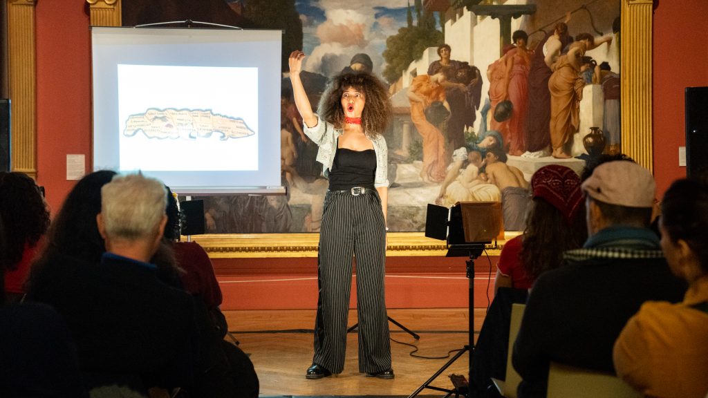 A woman of colour in front of a huge renaissance painting with an illuminated projection of a map, she has her arm raised over her head and is mid speech, her mouth a round o