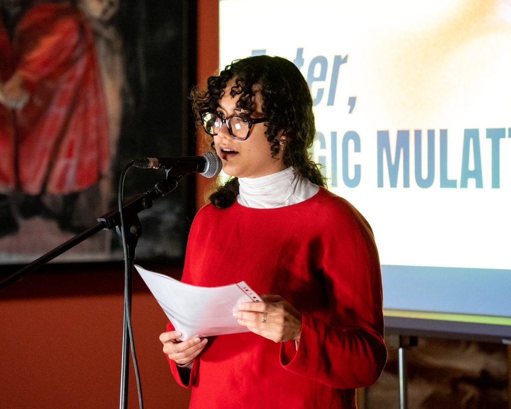 A mixed race women with dark curly hair in a red jumper stands behind a microphone holding a piece of paper