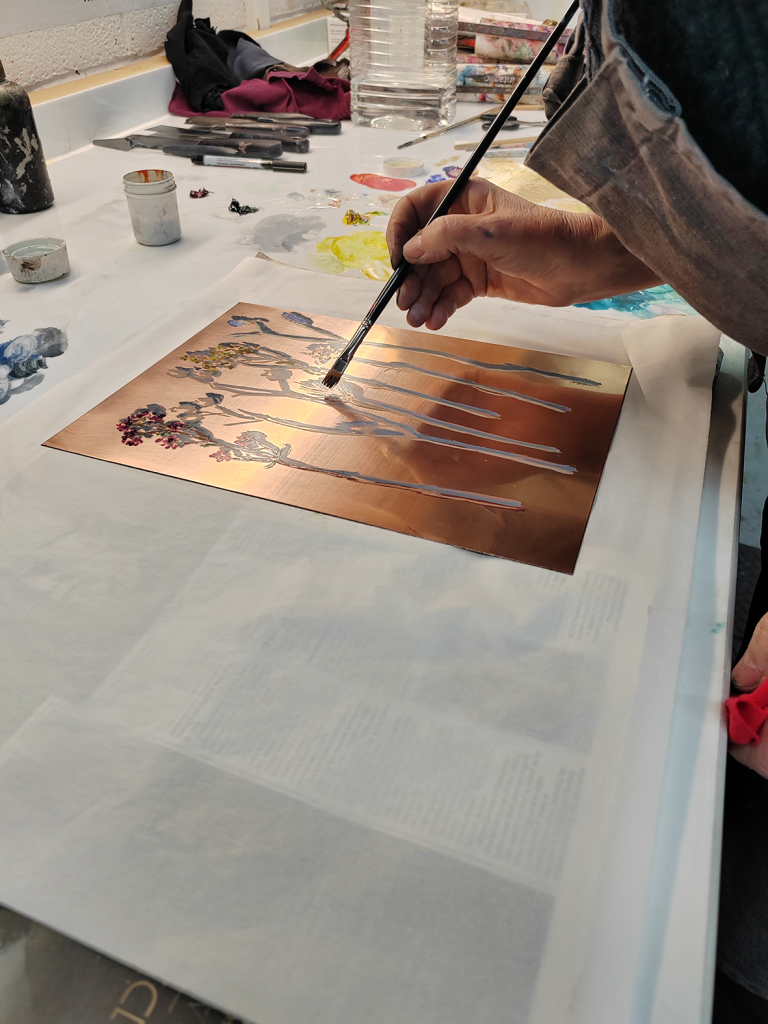 A golden coloured plate bearing images of flowers on long stalks, the artist leaning over with a brush in hand