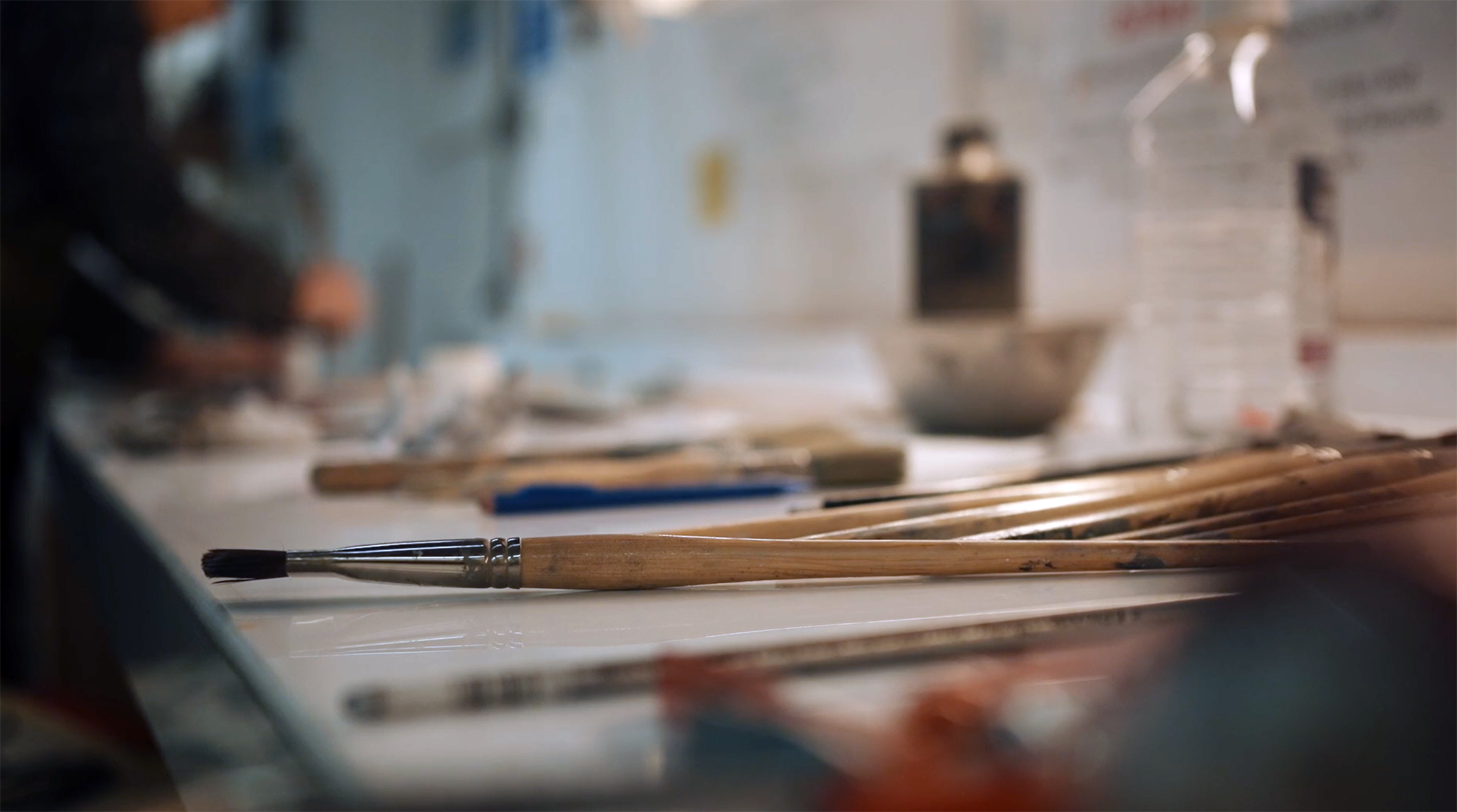 A close up shot of paintbrushes on a work bench with the artist blurry in the background