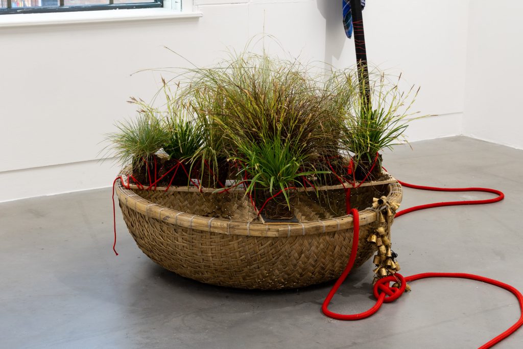 Another round basket-like coracle rests on the grey gallery floor. Grasses grow from within the basket, red ropes and golden bells spill out.