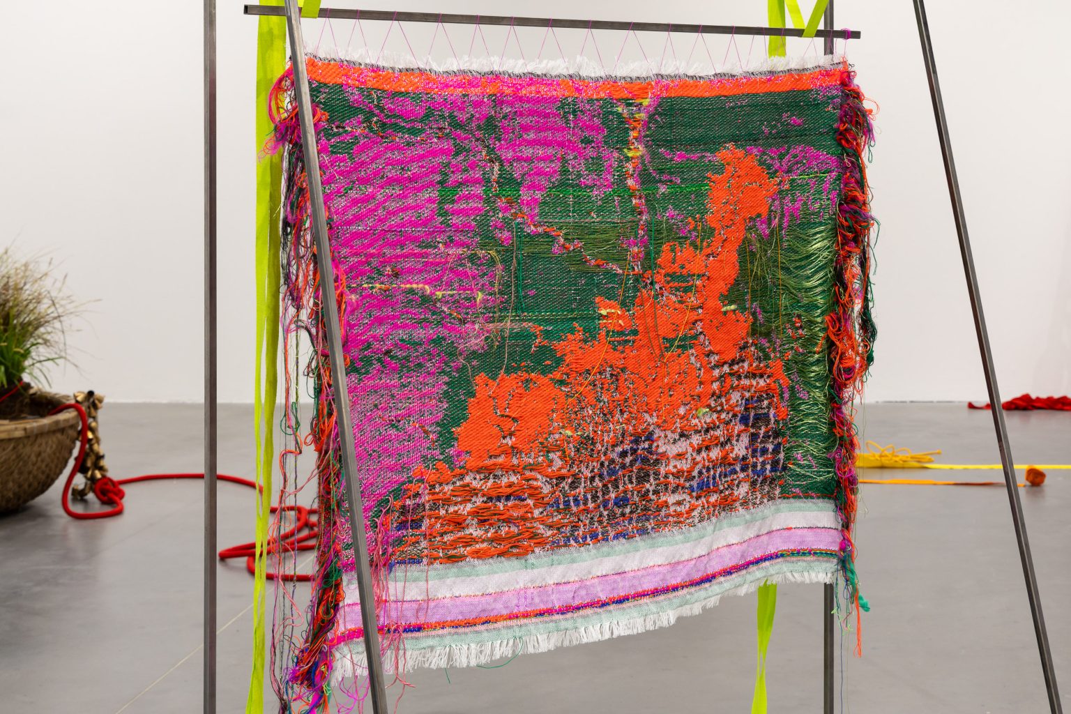 A woven textile piece is suspended from a metal frame in the centre of the gallery. Threads of green, bright reds and pinks form an abstracted image that perhaps resembles a disintegrating map of some kind. The edges of the weaving are left untrimmed and threads of different colours hang down.
