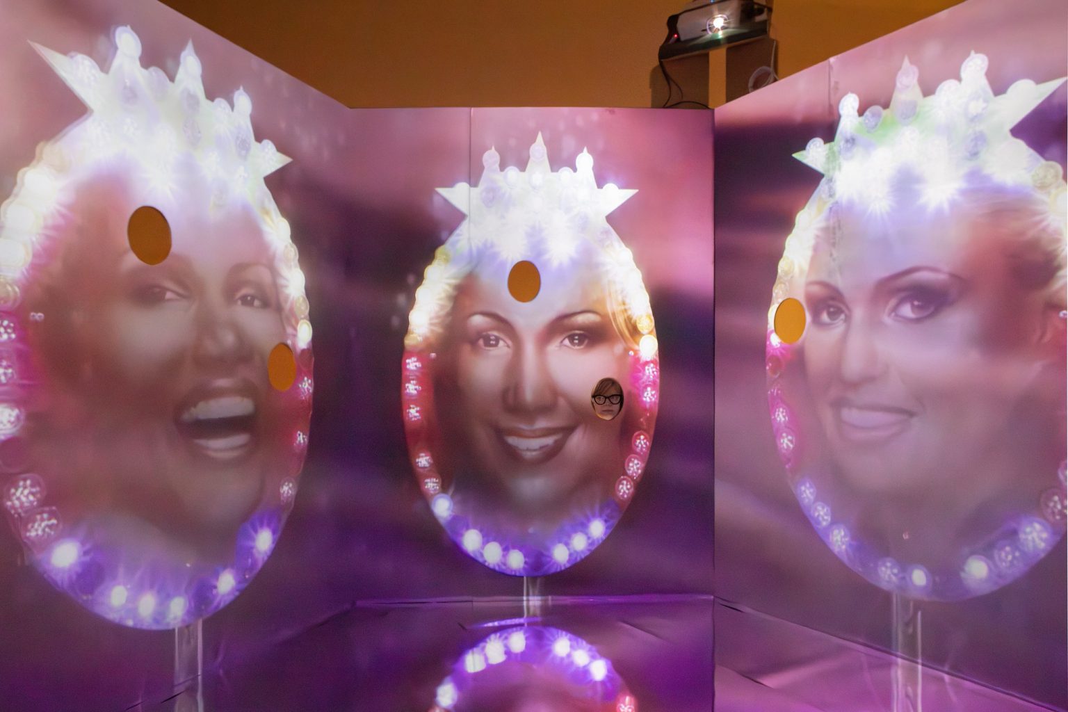 Three large female faces smiling against a purple backdrop with fairground lights. Holes are cut into the pictures for viewers to insert their own faces.