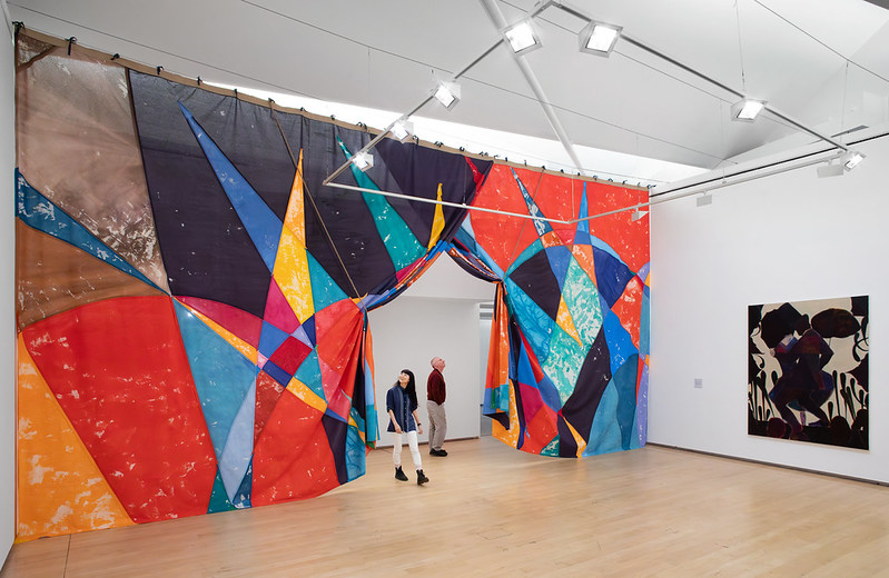 A large curtain cuts through a gallery space, with a red, blue, yellow and black triangles and circles pattern. Two people walk through an opening in the curtain, and a black and beige painting hangs on a wall to the right. 