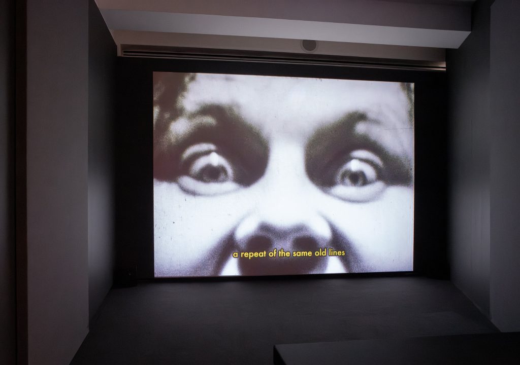 A darkened room with a large screen. The screen shows a close up of eye and a nose, a shocked expression. The yellow subtitles read "a repeat of the same old lines".