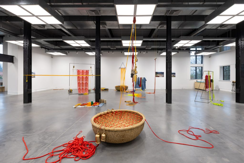 Installation view of Raisa Kabir 'I only dance, I wish we could sing' at Hub. In the foreground is a large circular basket with red rope and golden coloured bells. An installation of brightly coloured textile works and several wall-based artworks can be seen behind.