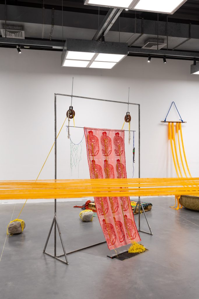 A textile banner is suspended from a black metal frame, which appears to be kept in place by bright saffron-yellow ropes tied to grey rocks on the gallery floor. A band of the same ropes cuts horizontally across the image in front of the artwork, apparently tied across the gallery space although the image is cropped in such a way to not reveal how or where they are secured.