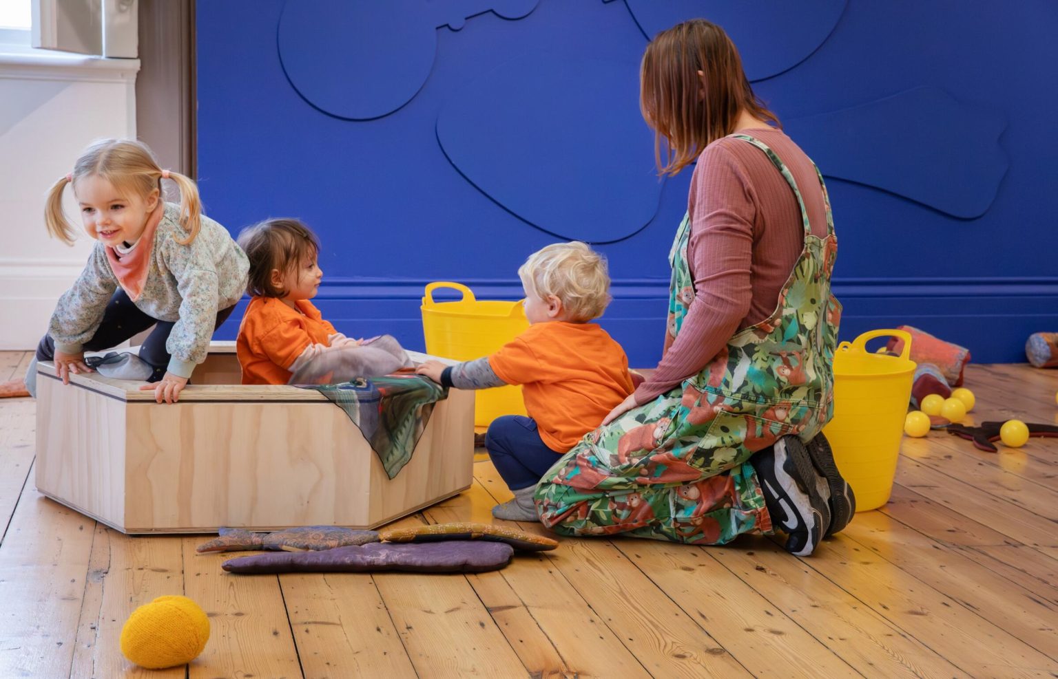 An adult kneels in the centre of the room beside a small wooden structure on which three toddlers clamber and play