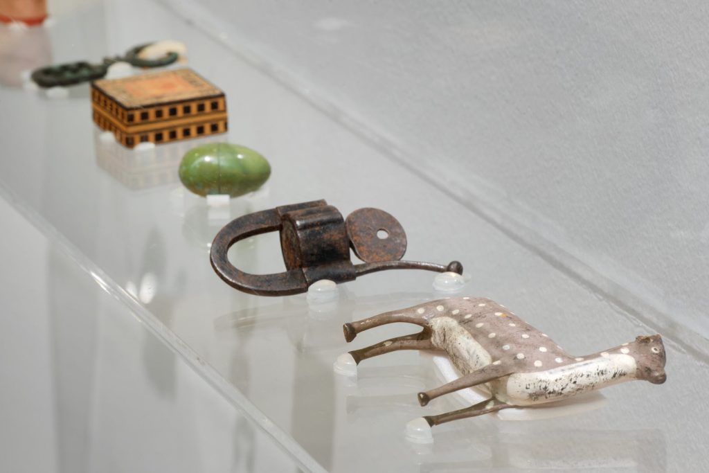 A closeup view of little objects on a clear perspex shelf including a statuette of a llama, a green egg, a small orange pill box and a rusty padlock and key
