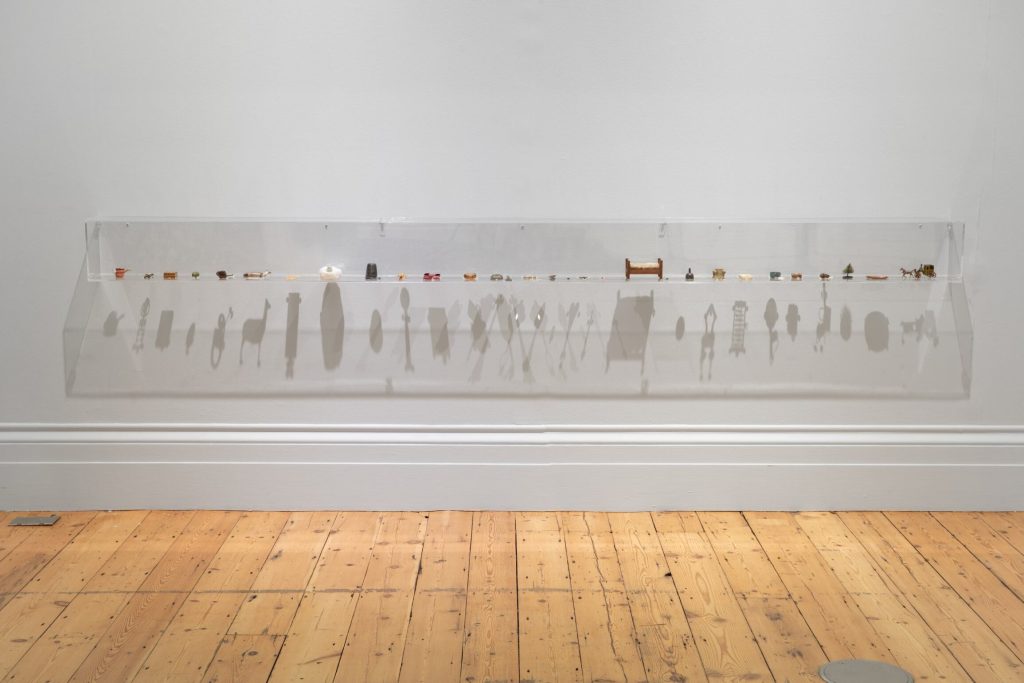 A white will with a clear perspex shelf running along it low to the ground with a single row of little trinkets and the shadows cast by them onto the wall from the lights above