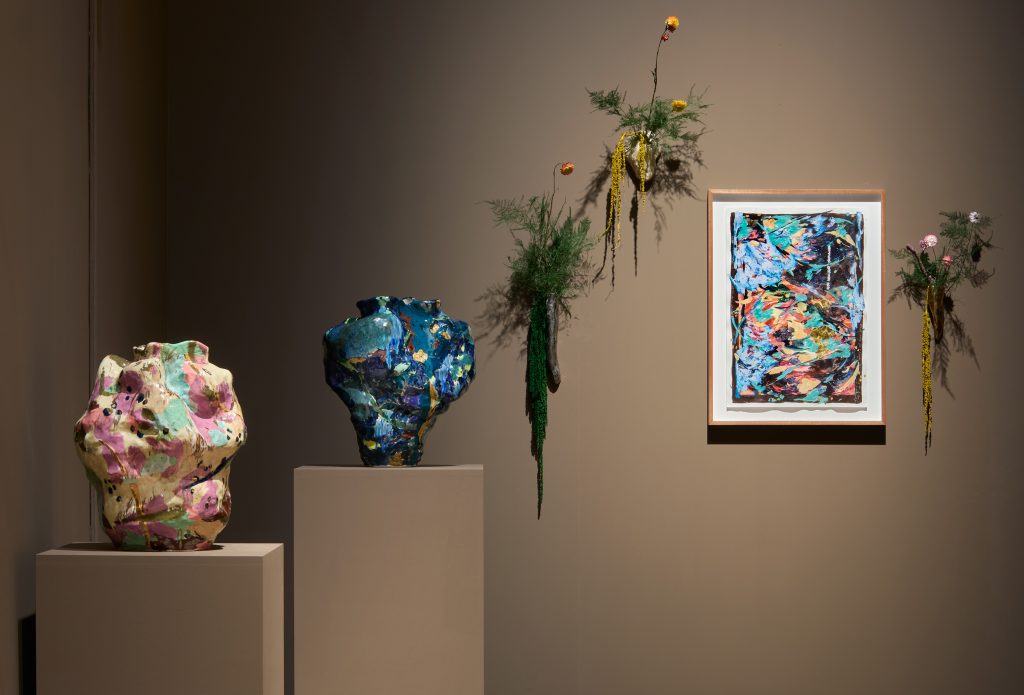 Two multicoloured lumpy ceramic vases on brown plinths beside a small, framed multicoloured abstract painting with plants wall decorations hung around it