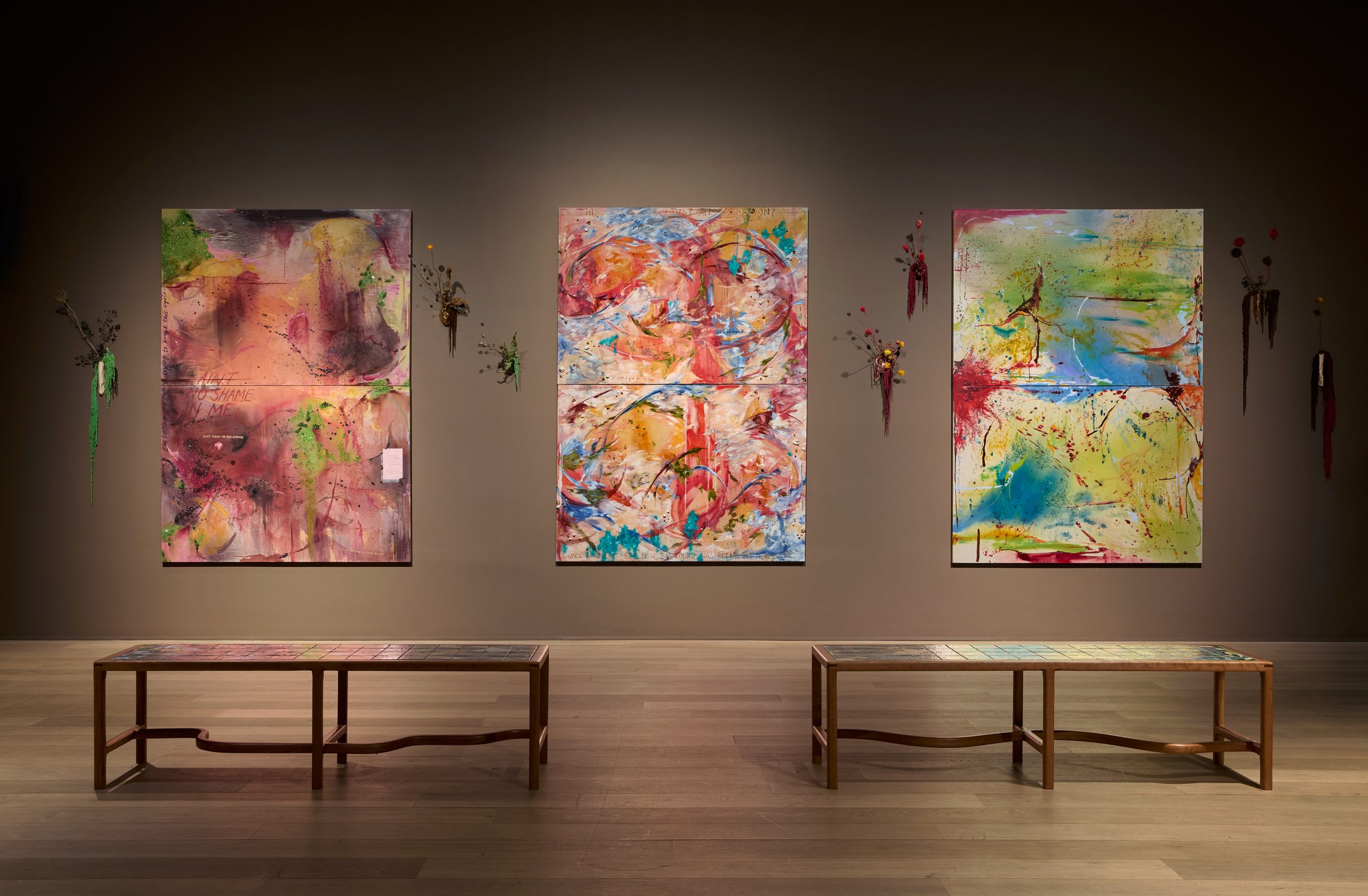 Three large, bright, multicoloured abstract paintings hung on a brown wall with two tile topped benches in front of them