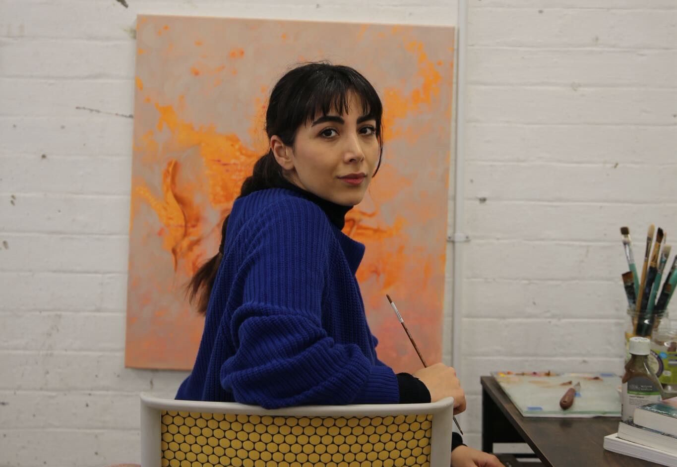 The artist with long hair in a ponytail and wearing a blue jumper turns away from a canvas to look into the camera