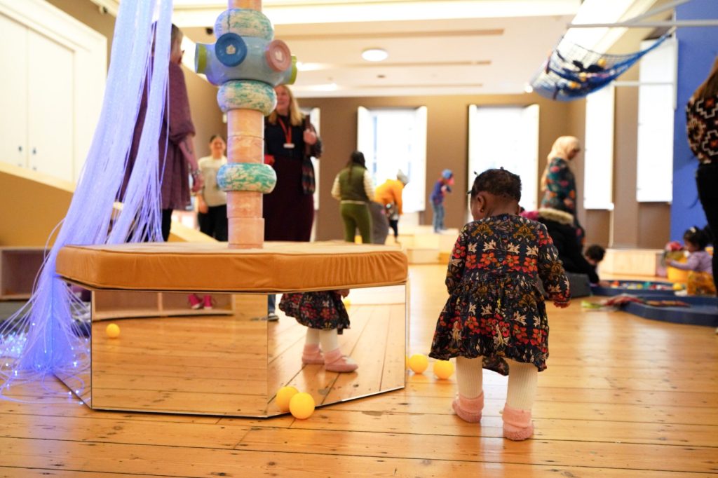 A toddler walking through the gallery space considering the reflection of their own feet in the low mirror they pass