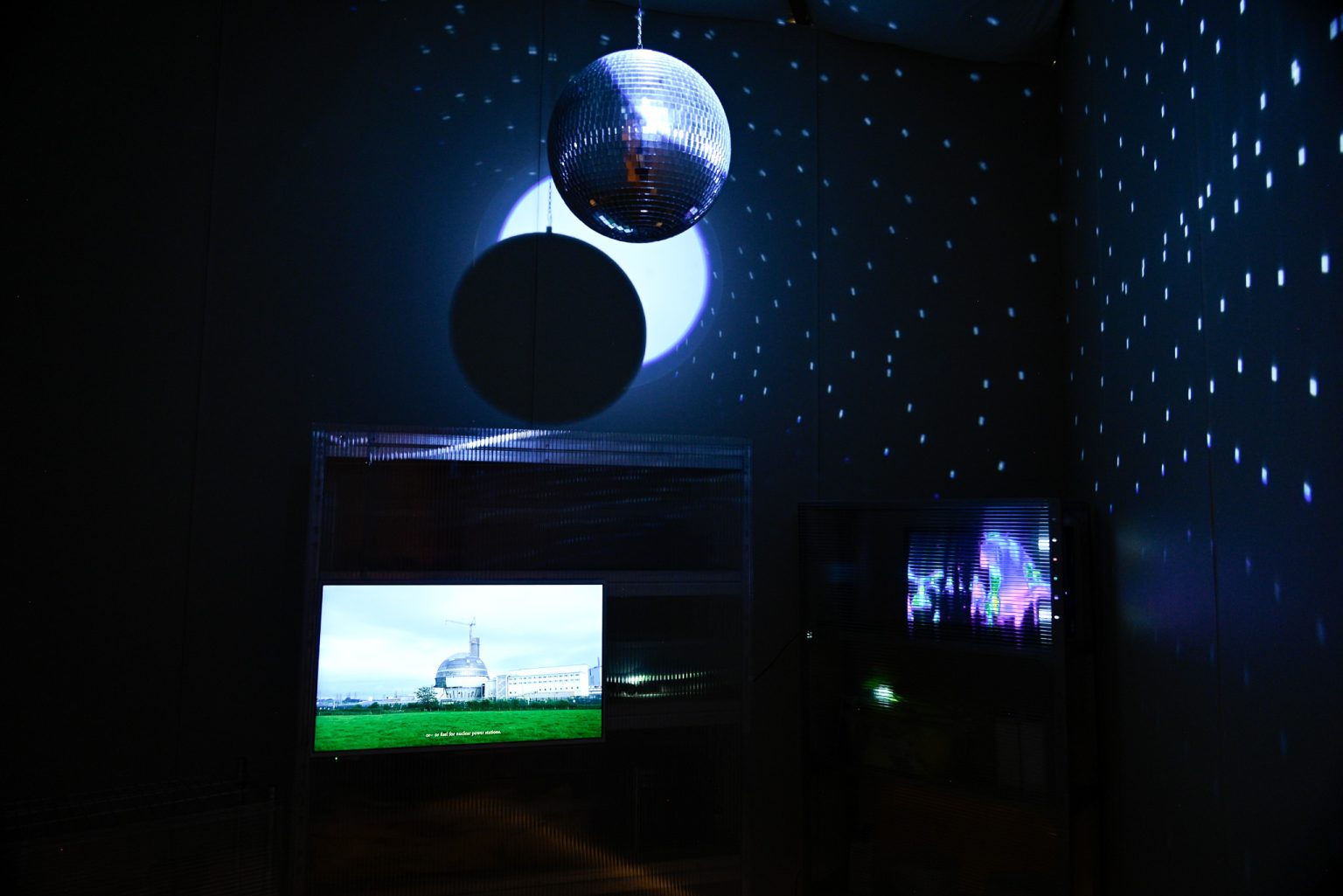 A dark room with an illuminated disco ball casting dots of light onto the walls above an illuminated screen showing futuristic buildings on the far side of a green lawn