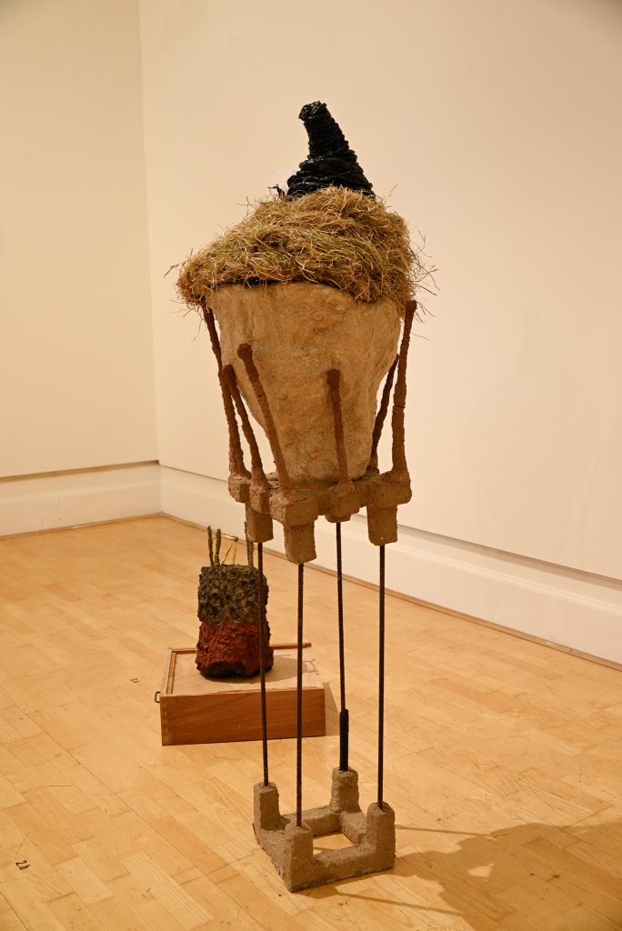 A tall sculpture of an earthlike clod with tufts of grass perched on very thin, spindly legs