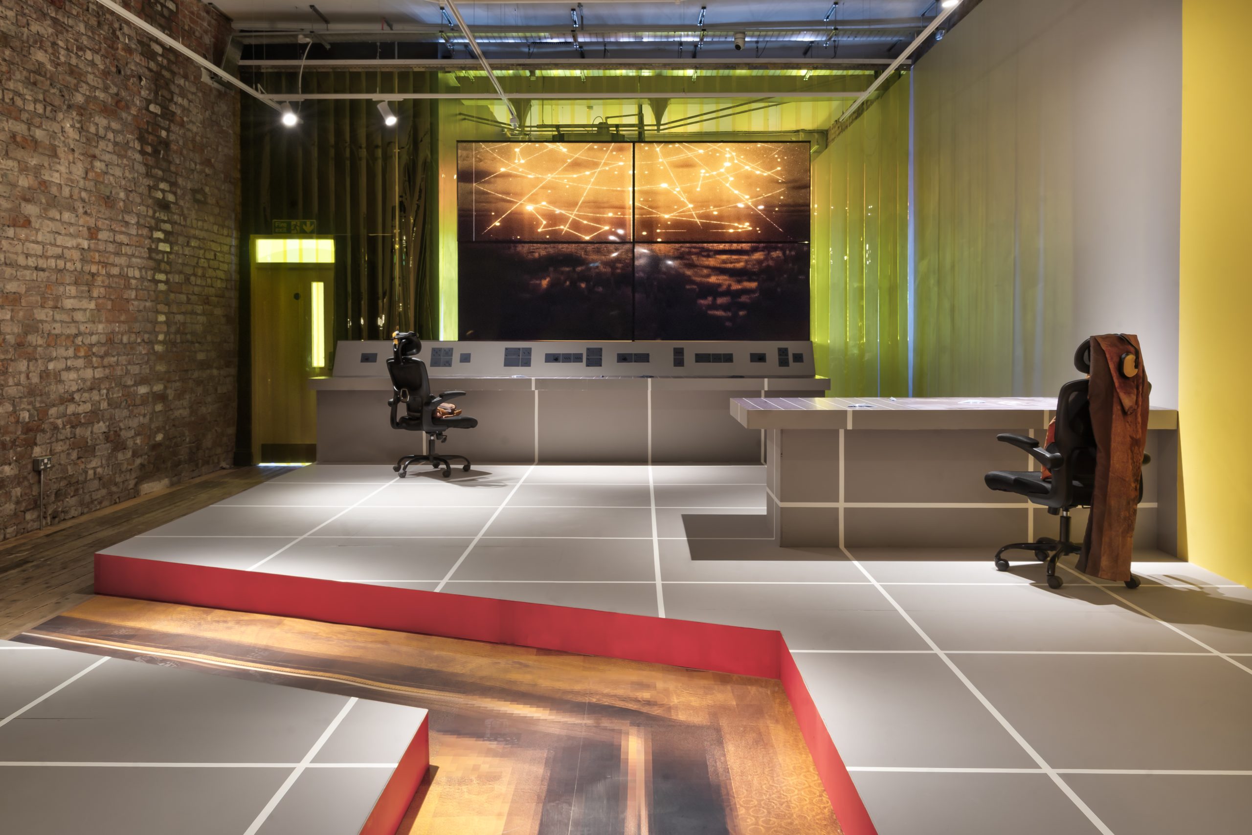 A grey floored room with a grey desk and grey control panel with knobs and dials underneath a huge screen with an illuminated night sky