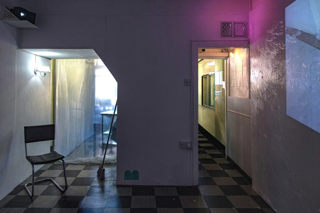 A dark room with two openings to a lighter room beyond - an ordinary doorway to the right, and a lower alcove-like opening covered in a transparent dust sheet and containing a chair and a light.