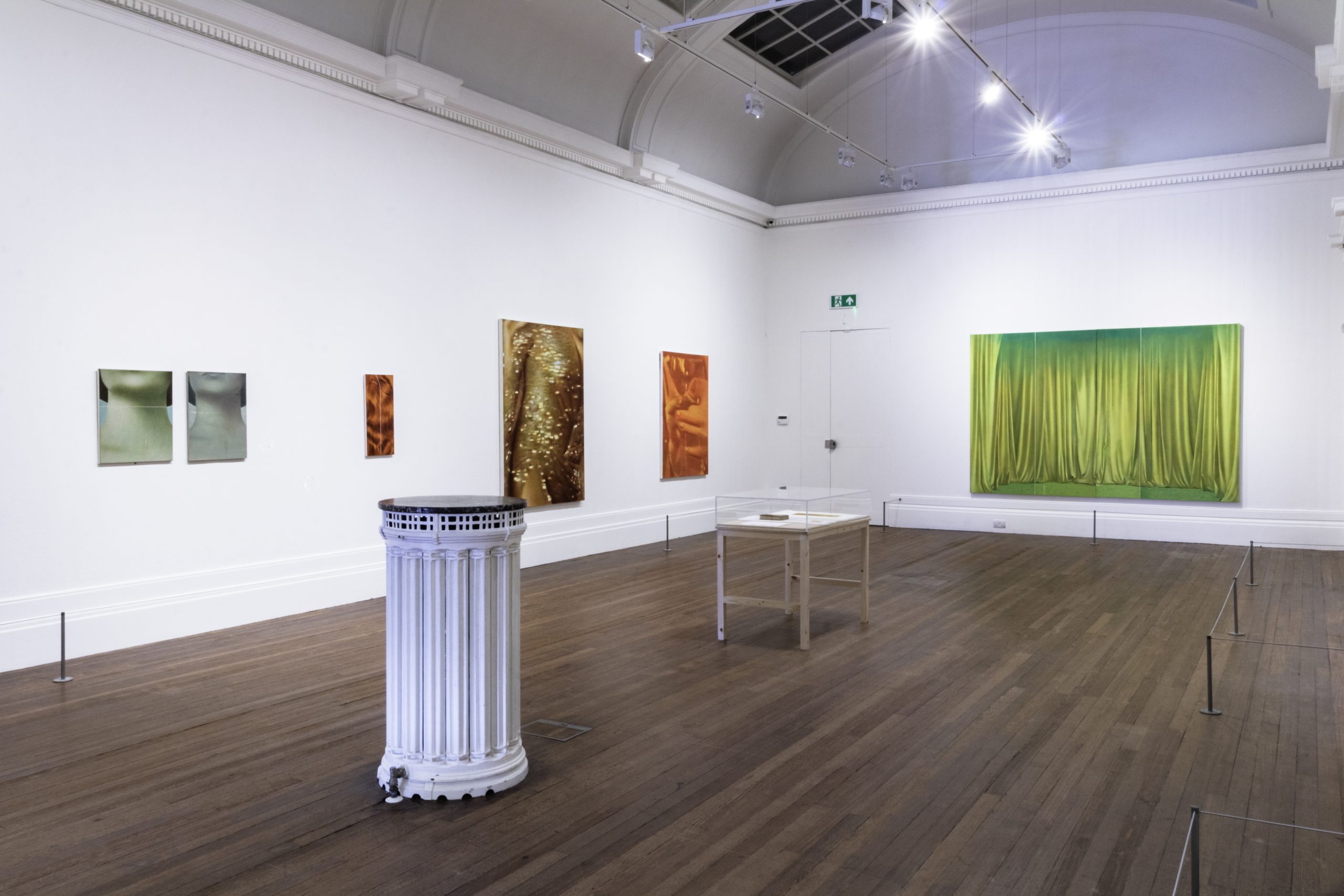 A wide view of the gallery, at the end of the room a painting of golden drapes with smaller paintings in browns and skin tones to the left