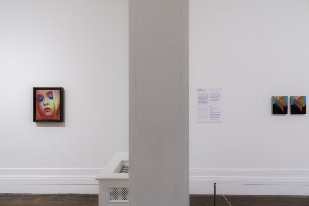 On the left a painting of a closeup of a young white girl's face with blue eyeshadow and red lips, with blonde hair falling around, and on the right two small paintings of a white man's brown beard against a blue background and his red shirt