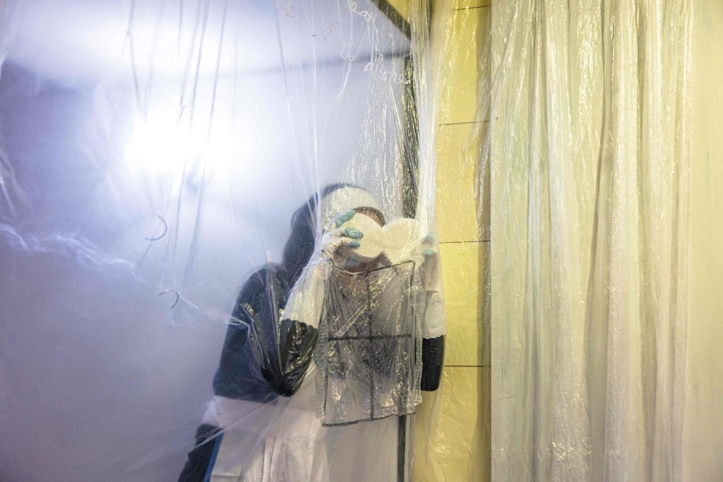 A person dressed as a maid holds a pair of plster eyes to her face and peers through a transparent dust sheet
