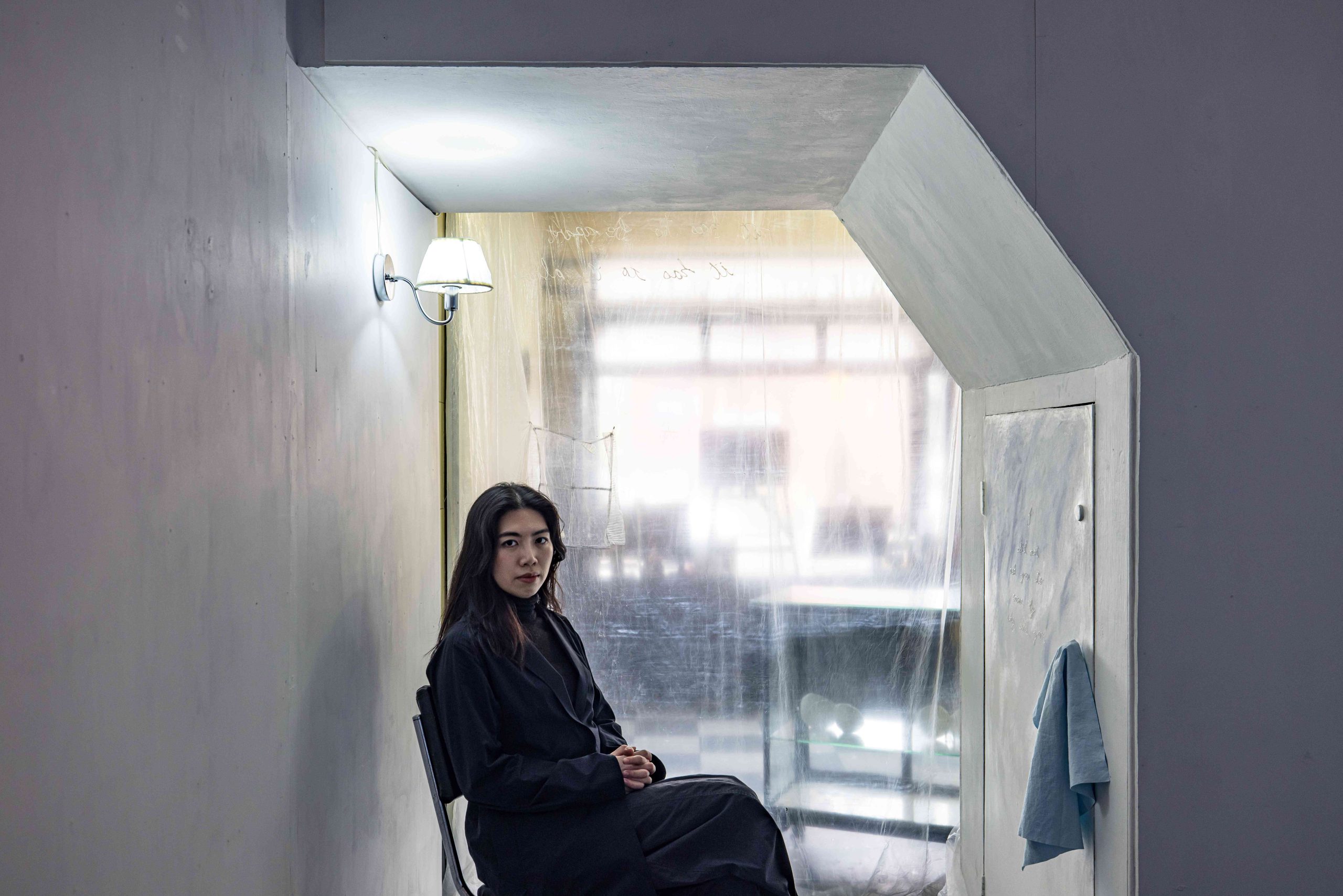 Photographic portrait of a person with long dark hair, dressed in dark clothing, seated in three-quarter pose, in an alcove below stairs, a brightly lit commercial space visible through a transparent curtain behind.