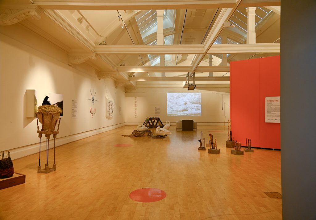 A wide view of the gallery in the roof of a building with white beams under the ceiling and artworks on the walls and floor