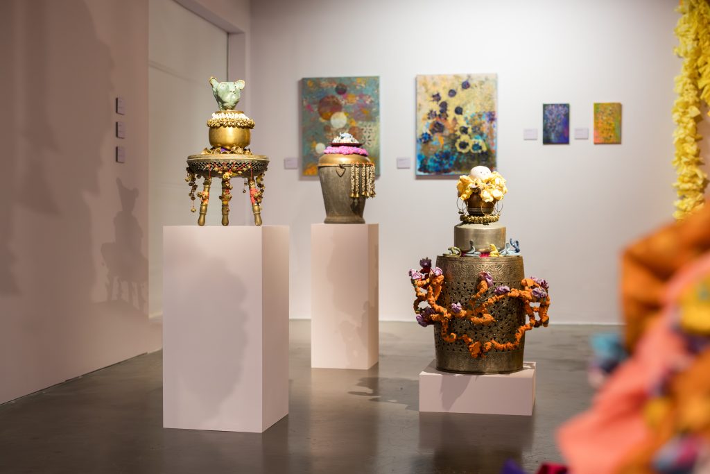 A wide installation photo showing metal vessels turned into sculptural objects and decorated with fabric, clay and other objects, placed on plinths. Behind the sculptures are four works on canvas that feature more muted colours applied in an expressionistic manner.