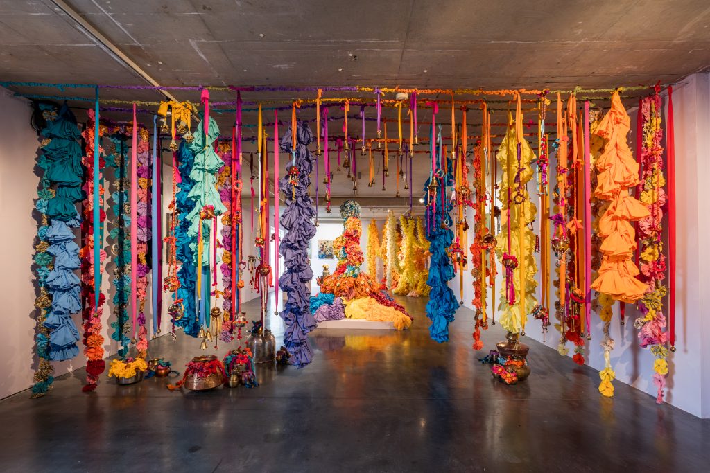A 'gate' of suspended fabric strips hangs aross the width of the gallery space. Small metal objects are attached to the fabric. There is a small opening in the artwork through which visitors can enter the exhibition.