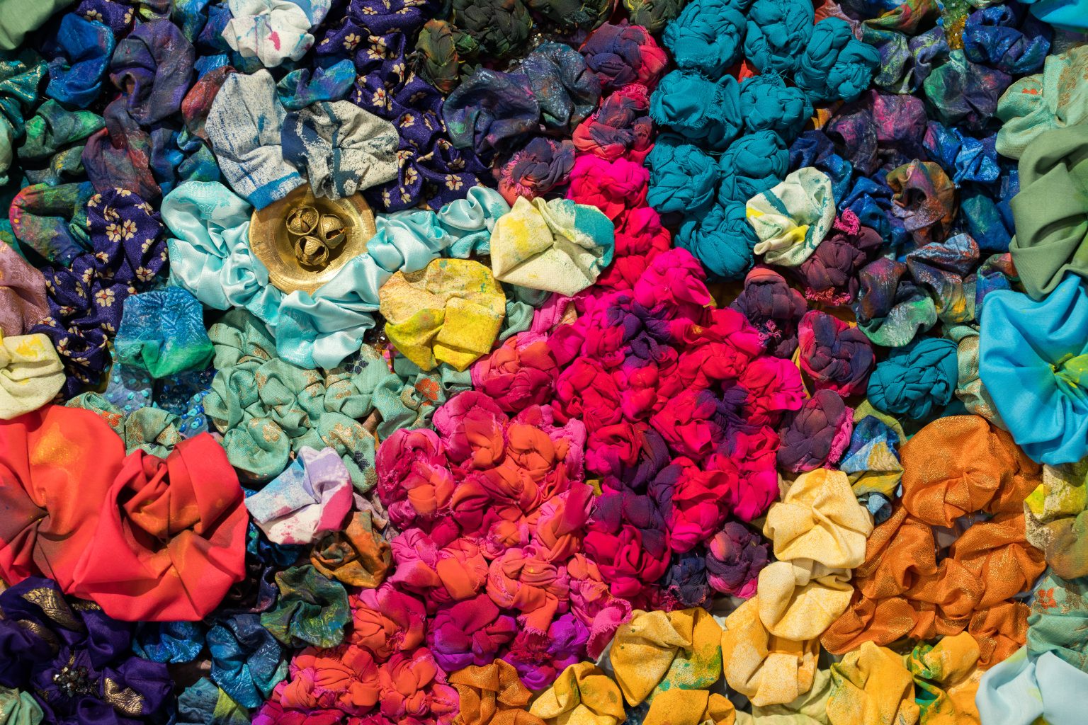 Detail of a textile artwork. Bright, multicoloured pieces of fabric have been knotted and bunched tightly together. A metal object with bells is pressed into the fabric.