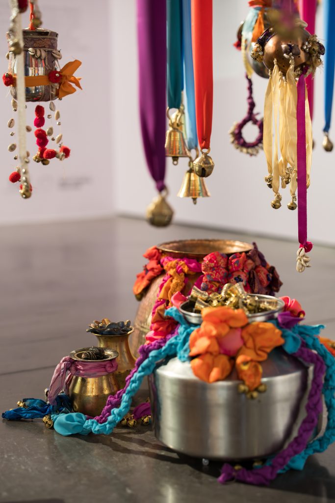 Metal vessels for carrying water are decorated with brightly coloured fabrics and placed in a grouping on the gallery floor. Bells and other objects are hung from colourful strips of fabric just above.