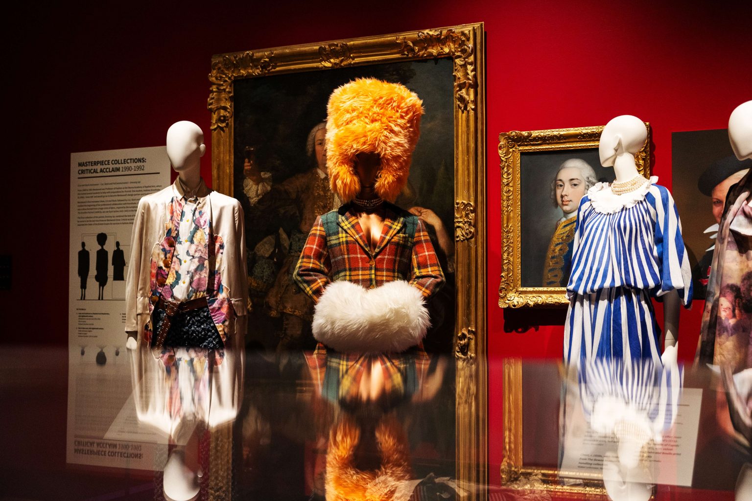 A red-walled, stately home gallery space. Three extravagant outfits on mannequins are visible across a reflective surface.