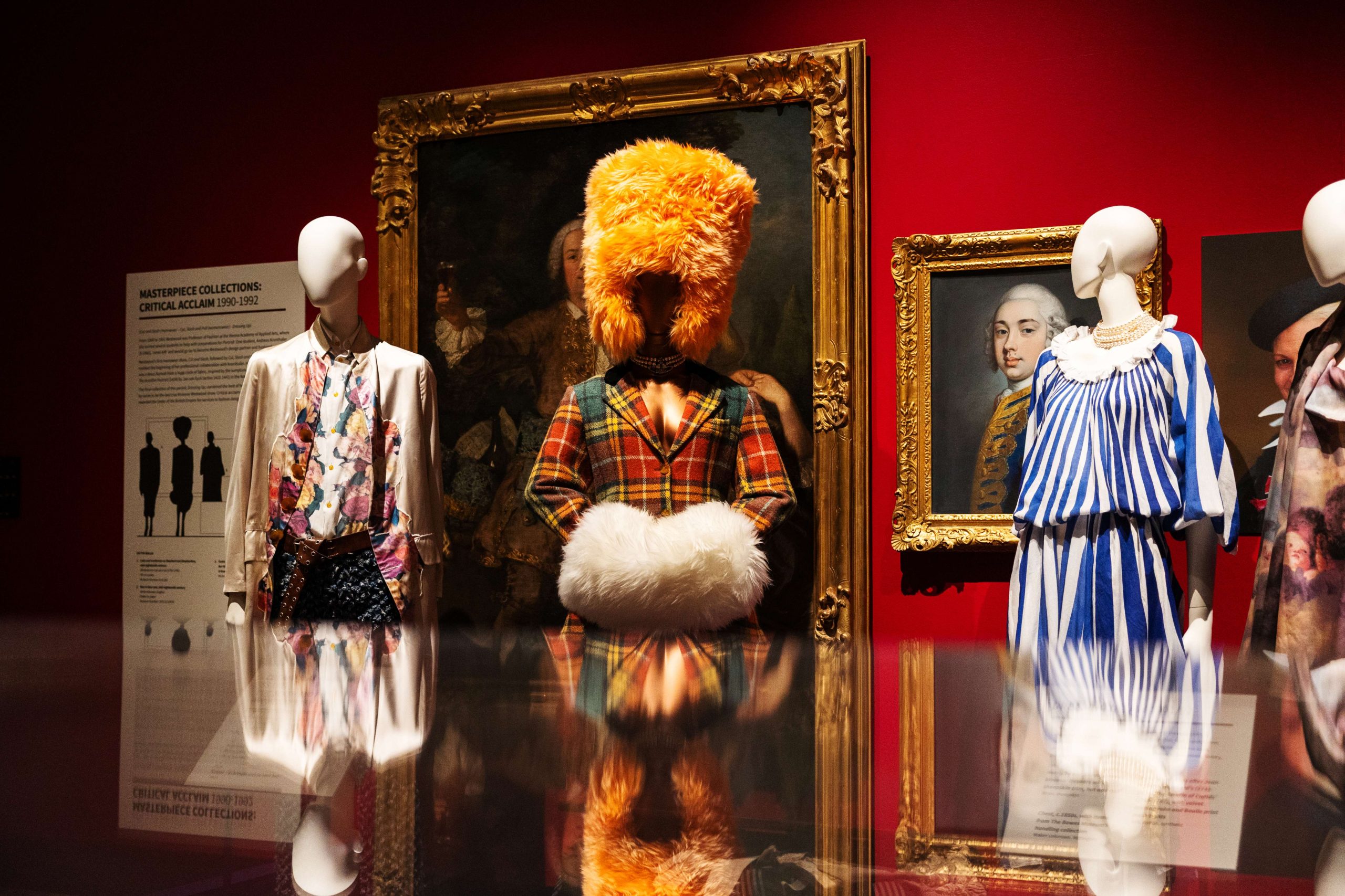 A red-walled, stately home gallery space. Three extravagant outfits on mannequins are visible across a reflective surface.