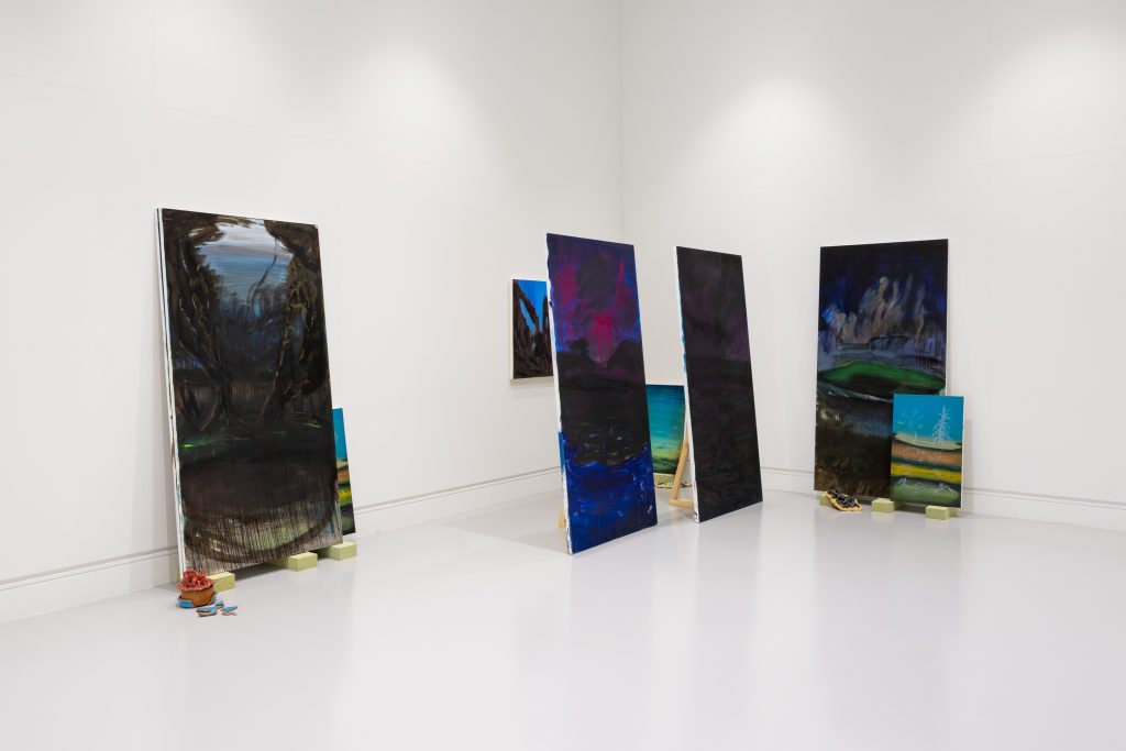 A series of large oil paintings in the corner of a gallery, propped on bricks and leant against the wall or wooden frames
