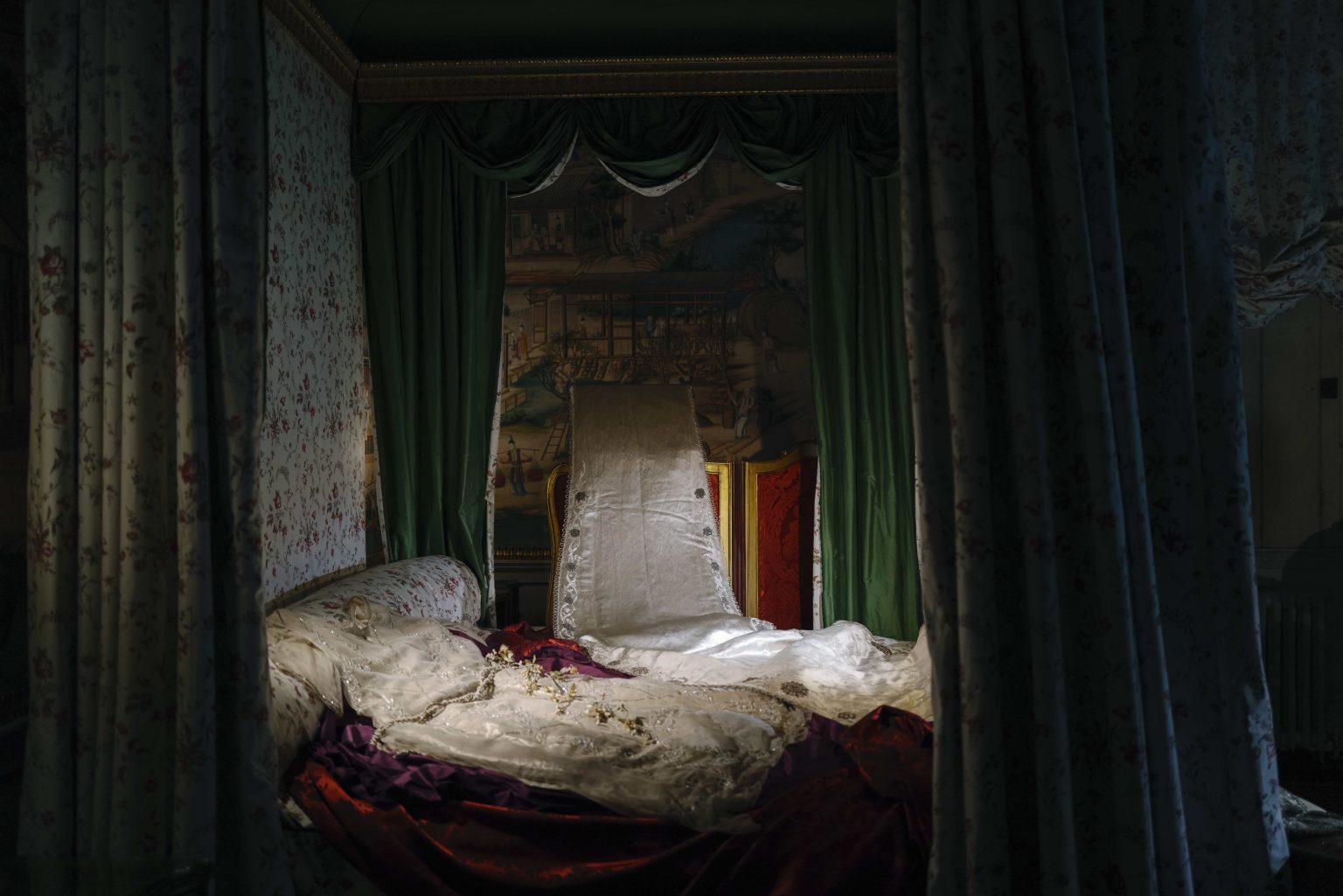 A lavishly made four poster bed, draped in rich fabrics and garments, in a dark, luxurious country house setting.