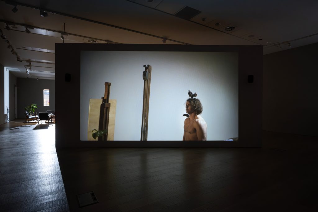 A man stands naked in an art school studio as if posing for a life drawing class. He is alone. Three starlings perch on his body; two on his head and one on his right shoulder.